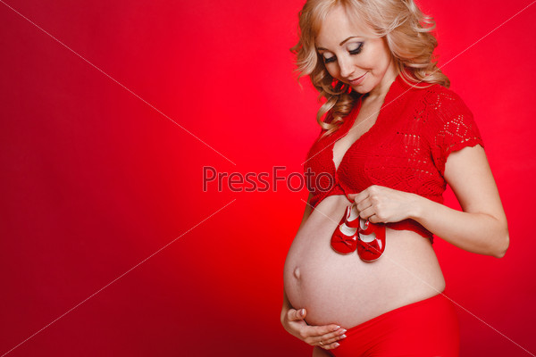 Portrait of Pregnant woman holding pair of red tiny shoes for baby on red background. Studio