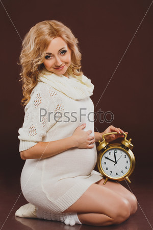 portrait of pregnant woman with clock in her hands on brown background. pregnant woman holding alarm clock. blonde young woman