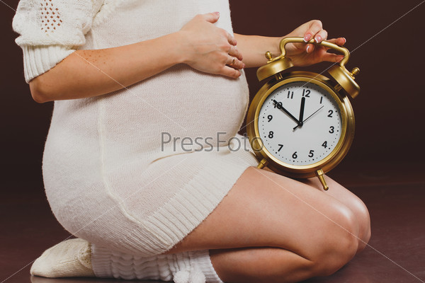 portrait of pregnant woman with clock in her hands sitting on brown background