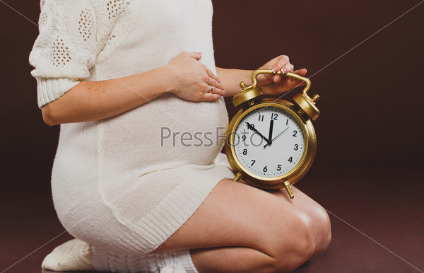 portrait of pregnant woman with clock in her hands sitting on brown background