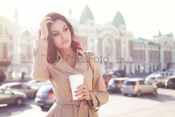 Beautiful young woman in a modern trench coat, holding a disposable takeaway cup and standing against urban city background.