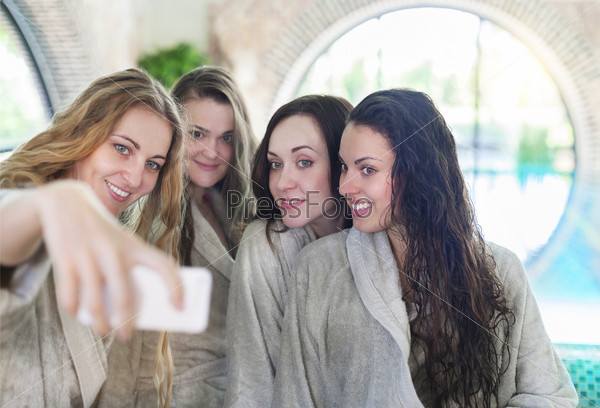 Four young women relaxing in the spa resort doing selfy wearing toweling robes