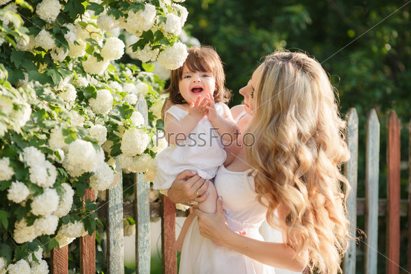 Mother with long blonde hair and flower wreath on had holding and playing with her baby girl in summer green park outdoor. Smiling mother and little daughter on nature. Happy people outdoors