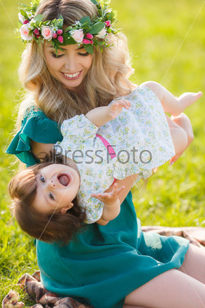 Mother with long blonde hair and flower wreath on had holding and playing with her baby girl in summer green park outdoor. Smiling mother and little daughter on nature. Happy people outdoors