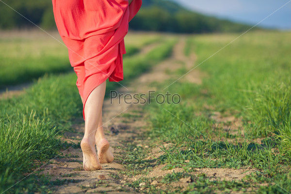 Portrait of barefoot woman in long red dress walking on the road in the green field in summer