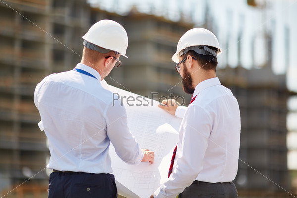 Young architects with blueprint discussing sketch of new building construction, stock photo