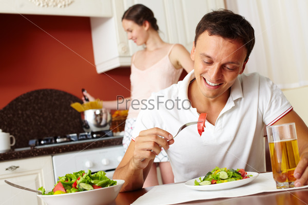 Happy man eating salad with his wife cooking on background