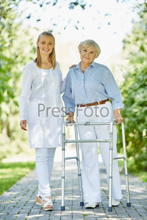 Smiling nurse and her aged patient with walking frame looking at camera outside