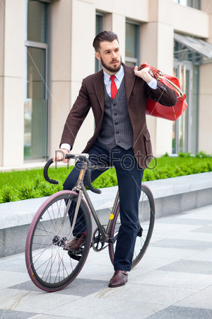 Handsome businessman in a jacket with red bag sitting on  his bicycle on city streets. The concept of the modern lifestyle of young men