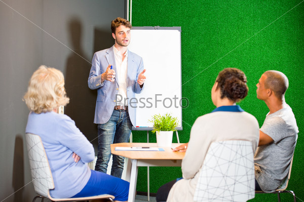 Young associate giving a presentation, using a flip-over white board and markers, to a small project team in an informal setting during a project team meeting in the lounge of a modern office
