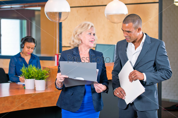 Two colleagues meeting in front of a reception desk where a secretary answers calls to discuss project notes