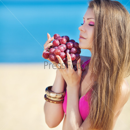 Beautiful woman with perfect hair and skin posing on the sea shore holding pink grapes. Summer picnic.