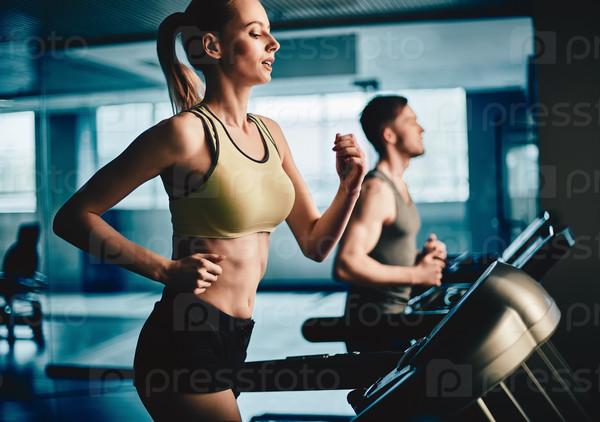Active young woman running on treadmill with guy on background, stock photo