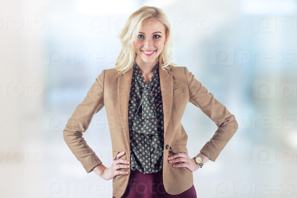 Bright young woman in brown jacket is standing in office with hands on her hips and smile brightly