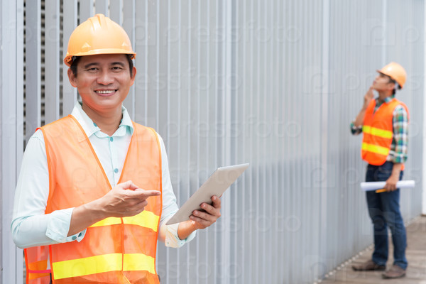 Cheerful Asian contractor pointing at the digital tablet in his hand