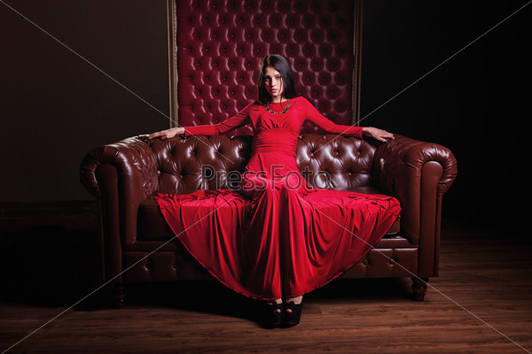 elegant sensual young brunette woman in red dress sitting on leather sofa and looking at camera