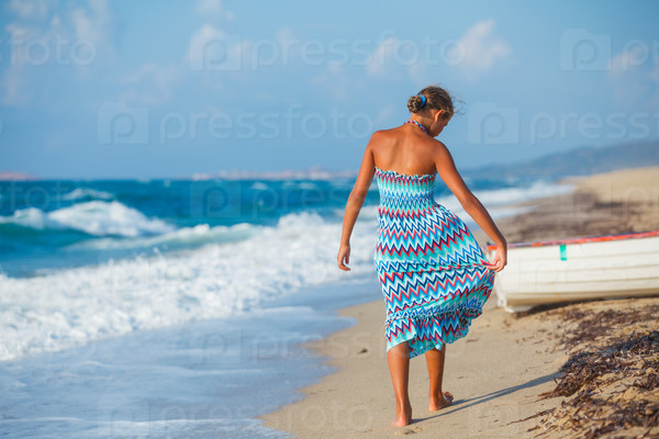 Young and beautiful girl in colorful dress walking on the beach near the ocean and looking far away at the sunset
