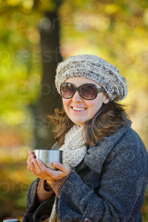 Young pretty woman with glasses and cap drinks tea in autumn park. Vertical view