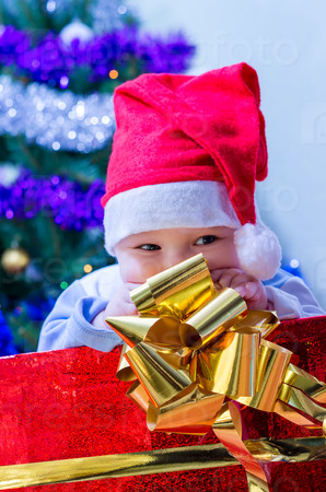 The kid sitting under the Christmas tree in a box with a gift from Santa Claus cap