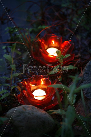 Glowing red candles by water, plants and stones