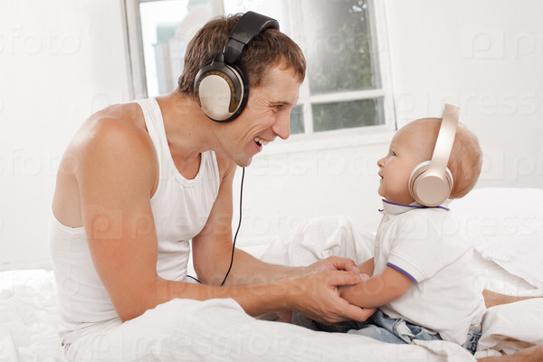 young father with his nine months old som on the bed at home on white background. Father and son wearing headphones