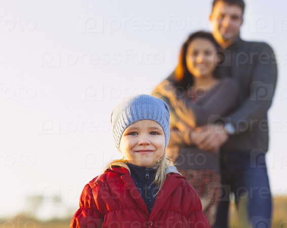 Portrait of the little girl with a funny hat outdoors and man and woman holding hands of smiling at the background. Family leisure outdoors concept