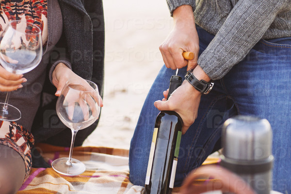 Young happy couple enjoying picnic on the beach together. Man opening bottle of wine. Close up