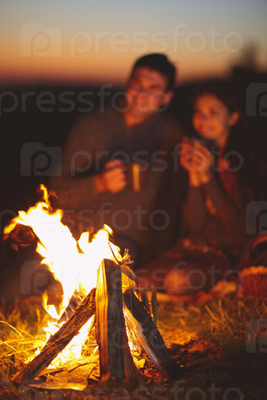 Portrait of the happy couple sitting by fire on autumn beach at the night. Focus on fire