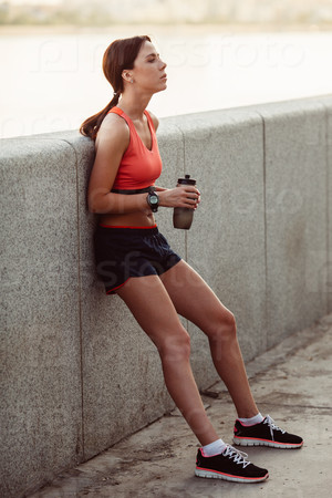 Female runner with bottled water tired from running standing near granite parapet