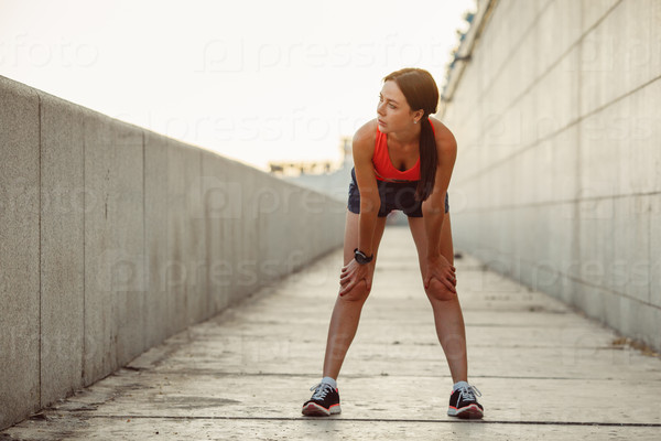 Young caucasian woman taking breath after jogging. Female athlete resting with hands on knees and looking away at sunset