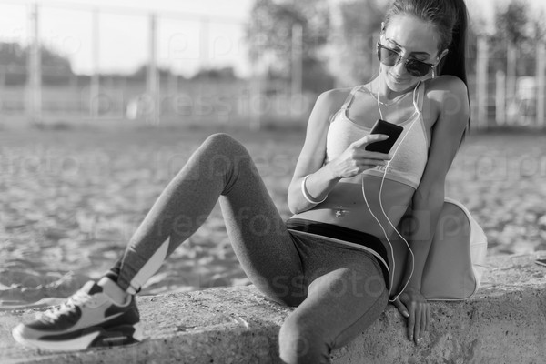 Beautiful fitness athlete woman wearing sunglasses resting listening music after work out exercising on summer evening in beach at sunset with smartphone outdoor portrait