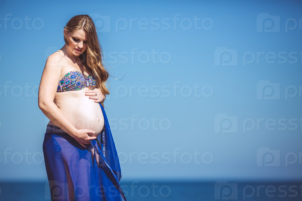 Portrait of pregnant beautiful woman on the seashore opposite blue sea and blue sky. Outdoors.