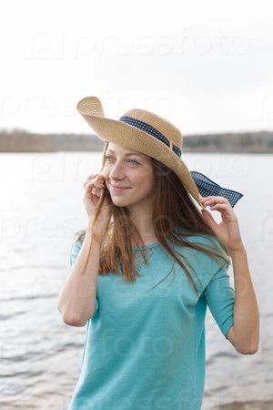Freckled girl in hat smiling and listening shell on the lake in windy weather