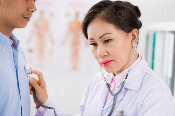 Mature Vietnamese doctor listening to heartbeat of her male patient