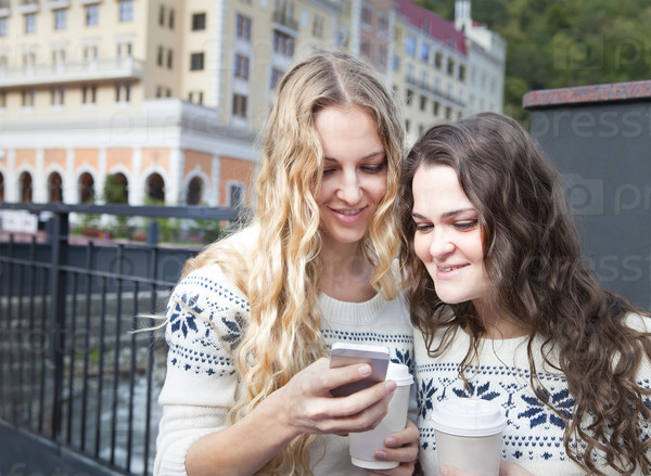 Two happy women friends sharing social media in a smart phone outdoors in a city. Two young women looking at mobile phone together while standing outdoors city. Drink coffee and discuss news