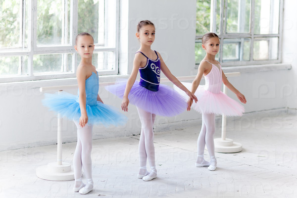 Three little ballet girls in multicolored tutu posing at ballet barre together on white background