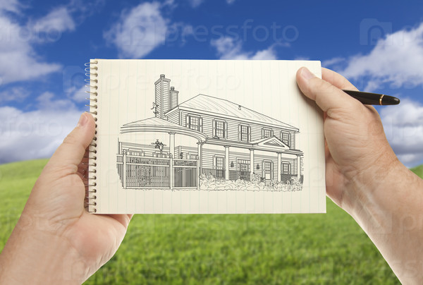Male Hands Holding Pen and Pad of Paper With House Drawing Over Empty Grass Field and Sky.