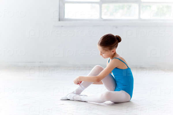 Beautiful little ballerina in blue dress for dancing sitting on the floor and puting on foot pointe shoes on white ballet studio background