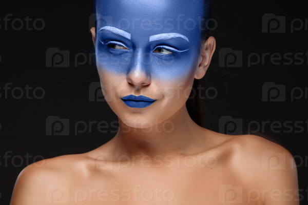 Portrait of a young woman who is posing covered with blue paint in the studio on a black background. Girl has blue lips and white eyebrows