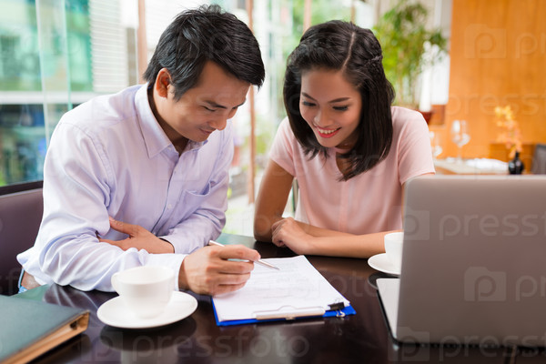 Asian smiling colleagues examining business contract while sitting in cafe