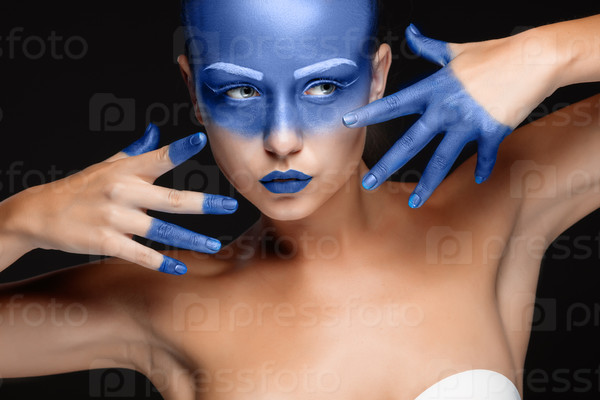Portrait of a young woman who is posing covered with blue paint in the studio on a black background. girl\'s hands around the face and painted blue