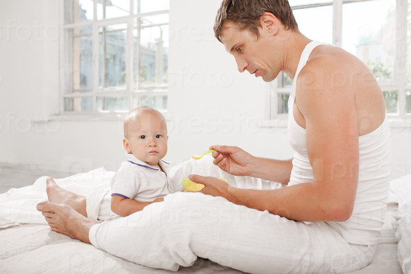 Young smiling father with his nine months old son on the bed at home on white home background. Dad feeding his son with a spoon