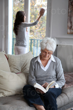 Elderly woman reading book while housekeeper cleans windows