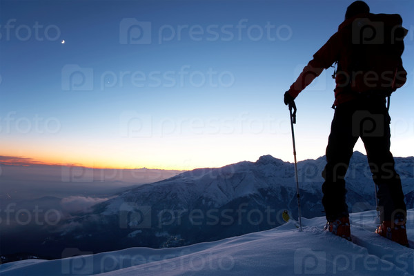 At dusk a brave backcountry skier reaching the summit of the mountain after a long day walking in the wilderness. Adventure and exploration concept.