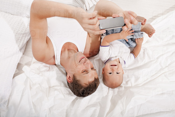 Young smiling father with his nine months old son  lying upside down on the bed at home on white home background. dad taking selfie photo with telephone camera