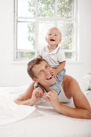 Young smiling father with his nine months old son on the bed at home on white home background. son sitting on his father\'s shoulders