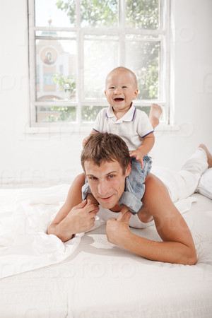Young smiling father with his nine months old son on the bed at home on white home background. son sitting on his father\'s shoulders