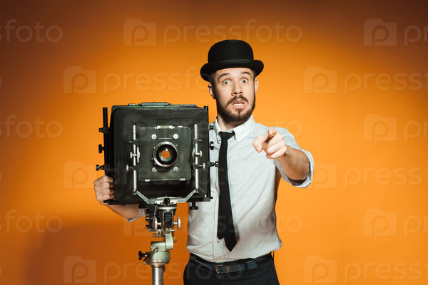 Young surprised man in hat as photographer with retro camera pointing to you on an orange background