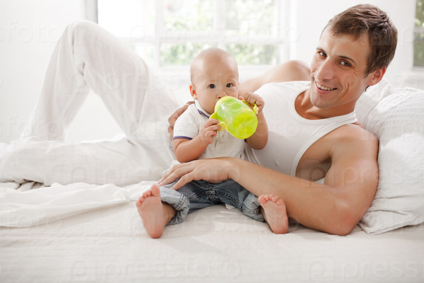 Young smiling father with his nine months old son on the bed at home on white home background. Child drinking water