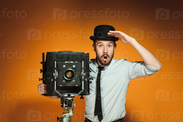 Young surprised man in hat as photographer with retro camera on an orange background